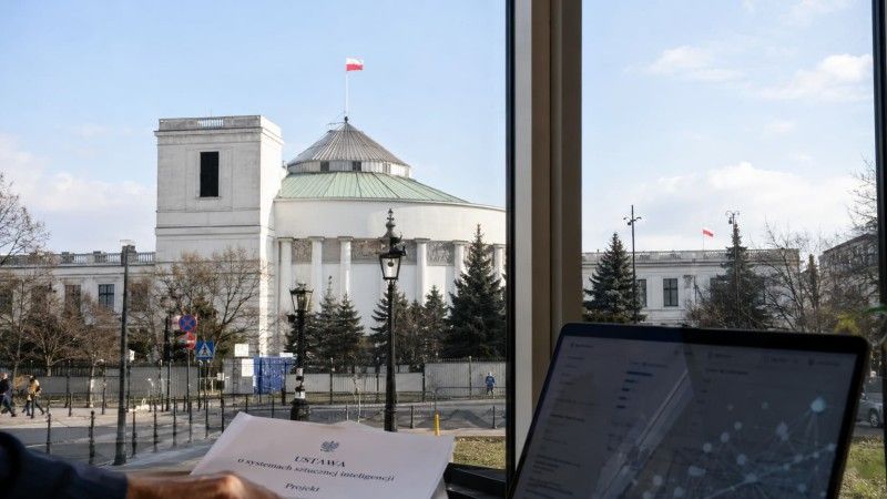 Budynek Sejmu RP w Warszawie widziany zza okna, na pierwszym planie dokument dotyczący ustawy o systemach sztucznej inteligencji oraz ekran komputera.