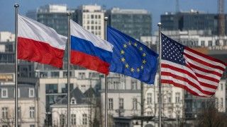 Cztery flagi — Polski, Rosji, Unii Europejskiej i USA — powiewają na tle nowoczesnej zabudowy miejskiej, ilustrując temat zagranicznych wpływów politycznych i sporów o finansowanie działań oddziałujących na debatę publiczną w Polsce.