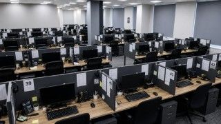 Puste, nowocześnie urządzone call center z dziesiątkami stanowisk pracy ustawionych w równych rzędach; na biurkach widać monitory, telefony, klawiatury i słuchawki.