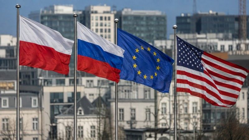 Cztery flagi — Polski, Rosji, Unii Europejskiej i USA — powiewają na tle nowoczesnej zabudowy miejskiej, ilustrując temat zagranicznych wpływów politycznych i sporów o finansowanie działań oddziałujących na debatę publiczną w Polsce.