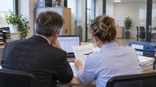 Kobieta i mężczyzna widziani od tyłu analizują dokumenty przy biurku z laptopem i segregatorami w jasnym biurze. Ilustracja wygenerowana przez AI.