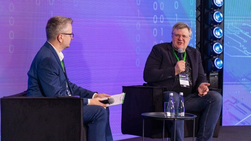 Sebastian Kondracki mówi do mikrofonu i gestykuluje podczas rozmowy z Piotrem Pilewskim na scenie Cyber24 DAYS, na tle fioletowo-niebieskiego ekranu z cyframi binarnymi.