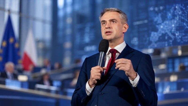 Krzysztof Gawkowski przemawia do mikrofonu na tle sali obrad w niebieskiej tonacji. W tle widoczne są rozmyte flagi Polski i Unii Europejskiej oraz ekran z mapą Europy i punktami danych.