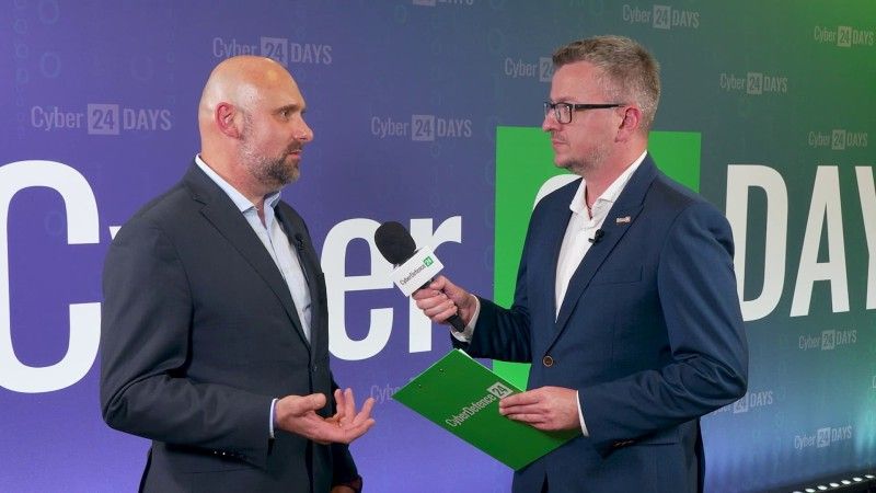 Tomasz Sawko z Orange Polska (po lewej) podczas rozmowy z Piotrem Pilewskim z CyberDefence24.pl na konferencji Cyber24 Days 2026. Obaj stoją na tle ścianki wydarzenia, a prowadzący trzyma mikrofon i zieloną teczkę.